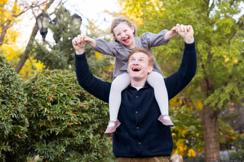 Fall photo session showing dad holding happy laughing child on his back in a park. 
