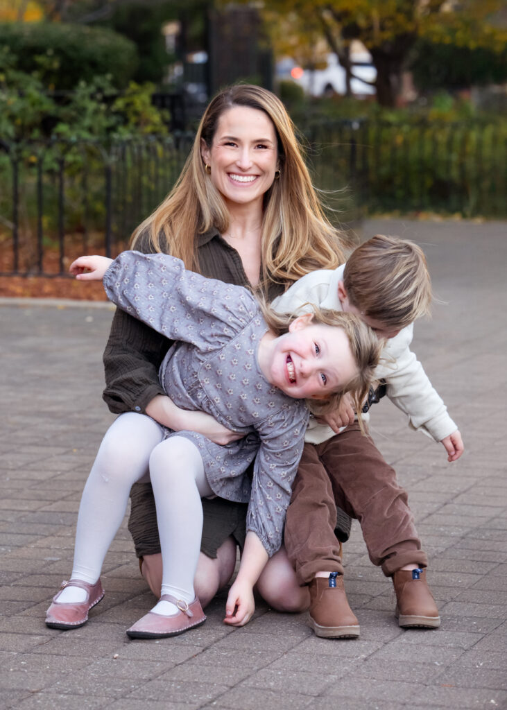 Mom smiling while holding her children that are playing and being silly during a family photo shoot. 
