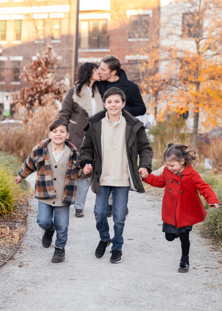 Parents kissing in the background whilde kids are running away in a family photo session in Hoboken.