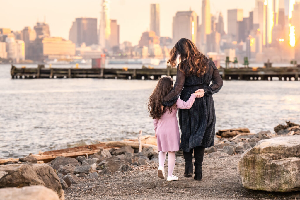 Mother and daughter walking hand in hand along the Hudson River at sunset with the Manhattan skyline glowing in the background during a family photography session.